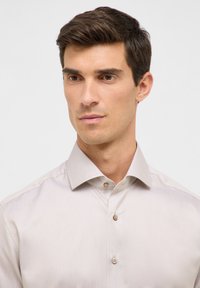 Camicia beige chiaro con un motivo testurizzato, dotata di un colletto strutturato e bottoni marroni sul davanti. Taglio dritto, design finemente aderente.