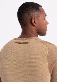 Maglione marrone a costine con una texture liscia, caratterizzato da un'etichetta marrone a contrasto sul retro del colletto e un design aderente.