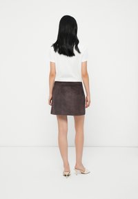 Donna con capelli neri lisci, che indossa una maglietta bianca a maniche corte, una mini gonna in suede marrone scuro e mule con tacco beige, in piedi di spalle.