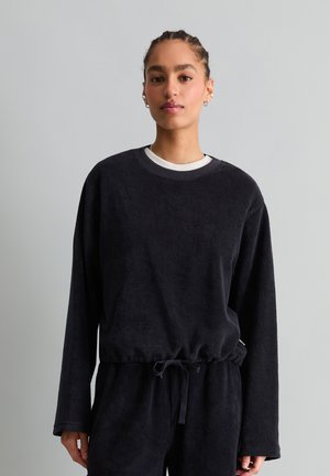 Jong persoon met gevlochten haar, gekleed in een zwarte trui met lange mouwen en bijpassende broek, staand tegen een effen grijze achtergrond.