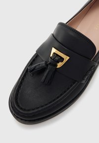 Mocassin en cuir noir avec une finition texturée, une semelle plate et un accent en métal doré avec des franges à l'avant. Design classique.