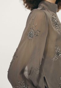 Gennemsigtig grå bluse med lange ærmer, der har detaljeret sølvfarvet edderkoppebroderi og perleaccent, høj krave og sidelommer.