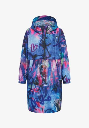 Femme portant une veste à capuche colorée et oversize avec des graffitis et des imprimés de chiens, jean retroussé, baskets jaunes, debout devant un fond blanc.