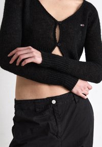 Cárdigan negro de punto con un escote en V profundo, dos botones y puños acanalados. Combinado con pantalones negros de cintura alta que tienen un botón de metal.