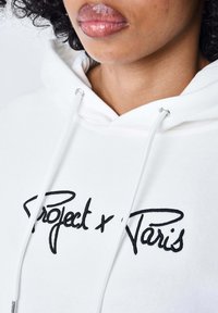 Felpa bianca con cappuccio e coulisse, con il testo ricamato in nero "Project x Paris". Materiale morbido con una vestibilità comoda, viso in primo piano con labbra lucide.