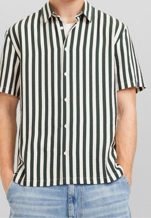 Mann trägt ein kurzärmeliges, geknöpftes Hemd mit vertikalen schwarzen und weißen Streifen, kombiniert mit hellblauen Jeans.