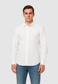 Camicia bianca a maniche lunghe con colletto button-down, bottoni frontali e dettaglio testuale verticale sul lato sinistro, abbinata a jeans blu aderenti.