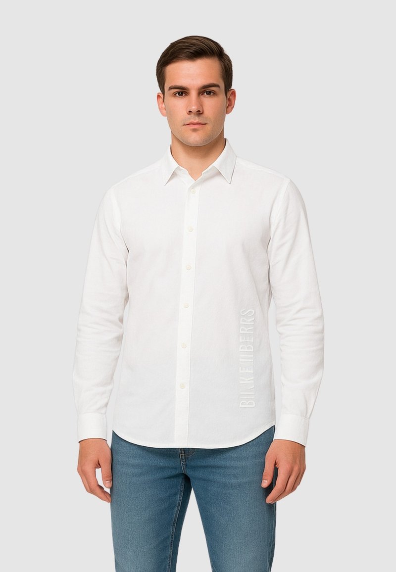 Camicia bianca a maniche lunghe con colletto button-down, bottoni frontali e dettaglio testuale verticale sul lato sinistro, abbinata a jeans blu aderenti.