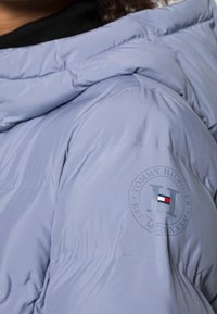 Zbliżenie na rękaw jasnoniebieskiej kurtki puchowej z okrągłą łatą z logo Tommy Hilfiger.