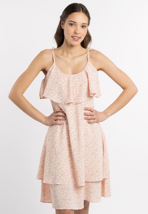 Jeune femme aux longs cheveux foncés porte une robe rose à fleurs sans manches à plusieurs couches avec de fines bretelles, debout les mains sur les hanches.