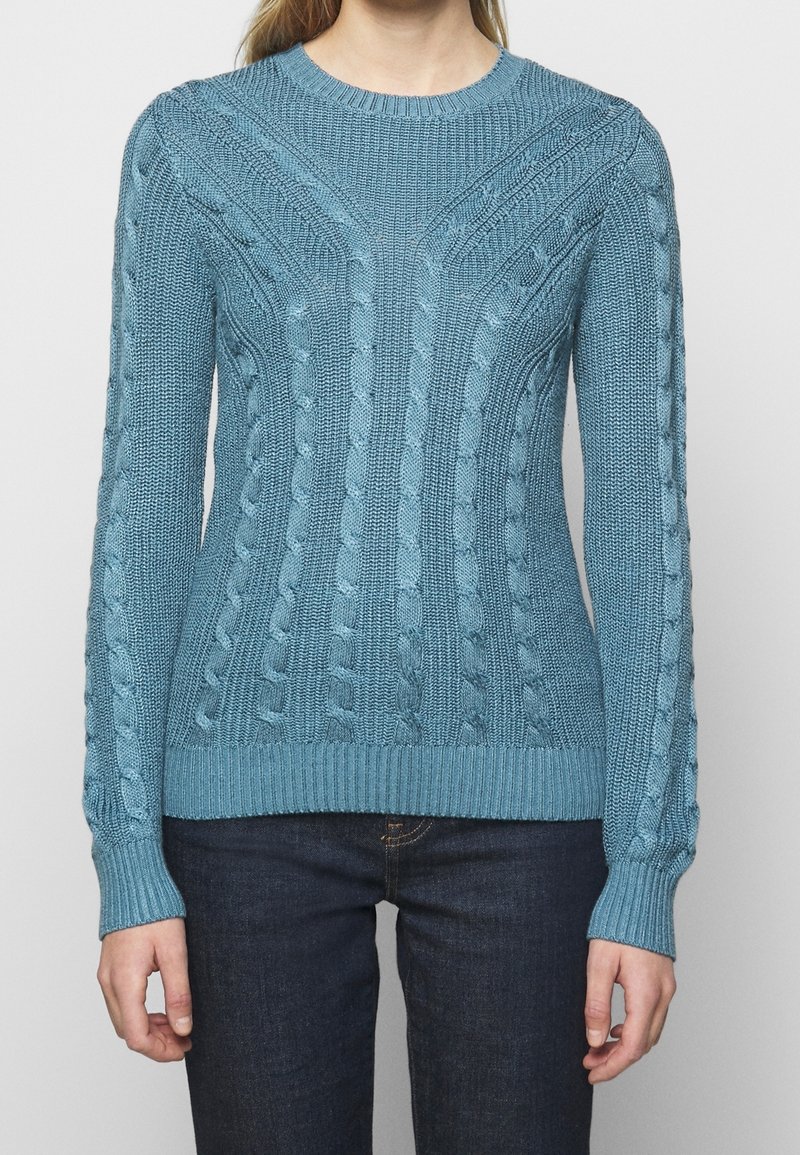 Femme portant un pull ajusté bleu clair à torsades, à manches longues et col rond, associé à un jean bleu foncé.
