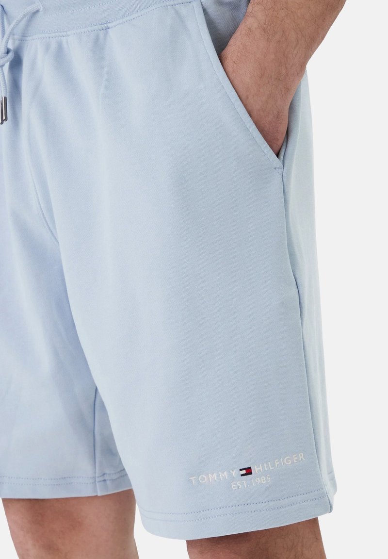 Shorts bleu clair avec poches latérales, une main dans une poche, et le logo "Tommy Hilfiger EST. 1985" près de l'ourlet.