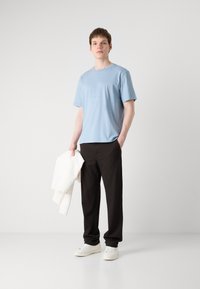 Lys blå crew-neck t-skjorte, bomullsmateriale, svarte straight-fit bukser, hvit hettegenser hengende over armen, og hvite joggesko.
