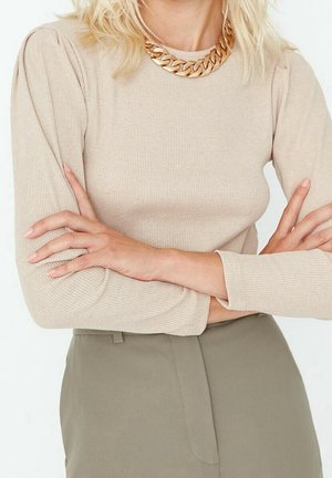 Frau mit verschränkten Armen, die ein beige geripptes Langarmshirt, grüne Hosen und eine dicke goldene Gliederkette trägt.