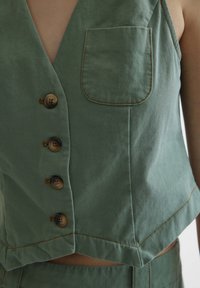 Grønn avkortet vest laget av teksturert stoff med fem brune knapper, en frontlomme og kontrasterende sømdetaljer.