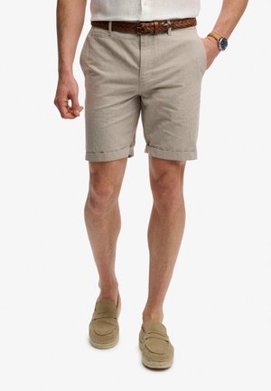 Mann trägt beige hochgekrempelte Shorts mit geflochtenem braunem Gürtel, beige Loafers, weißes Hemd und Armbanduhr, steht mit einer Hand in der Tasche.