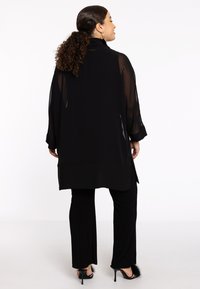 Kimono transparent noir à manches longues, superposé sur un pantalon ajusté assorti, présentant un ourlet long et des accents texturés sur les chaussures.