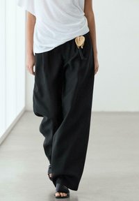 Persona che indossa pantaloni neri larghi con una tasca intrecciata con coulisse in vita e sandali neri aperti sulle dita, camminando al chiuso.