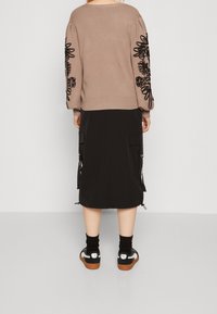 Pull tricoté couleur fauve avec broderies florales noires sur les manches, assorti à une jupe midi noire avec poches. Baskets noires et grises.