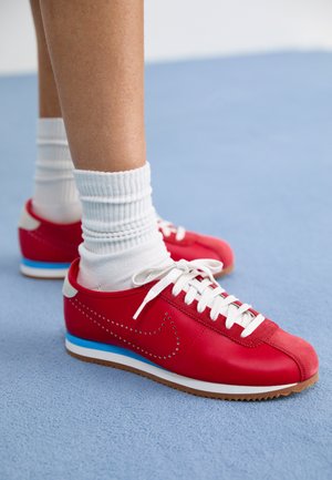 Piros bőr sportcipő fehér cipőfűzőkkel és perforált díszítéssel, kék középtalppal és bézs sarkakkal, fehér bordázott zoknival párosítva.