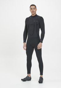 Schwarzer Radanzug mit langen Ärmeln und Trägern, ausgestattet mit einem hohen Kragen, taillierter Passform und strukturiertem Stoff, kombiniert mit schwarzen Radsportschuhen.