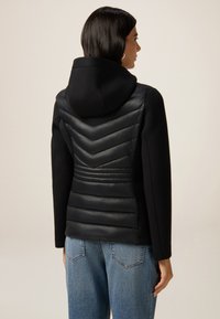 Frau trägt eine schwarze Kapuzenjacke mit gestepptem Zickzack- und horizontalem Polsterdesign auf dem Rücken, kombiniert mit blauen Jeans.