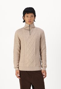 Pull en tricot beige avec col montant, fermeture éclair, motifs texturés avec câbles et cercles, et poignets et ourlet côtelés.
