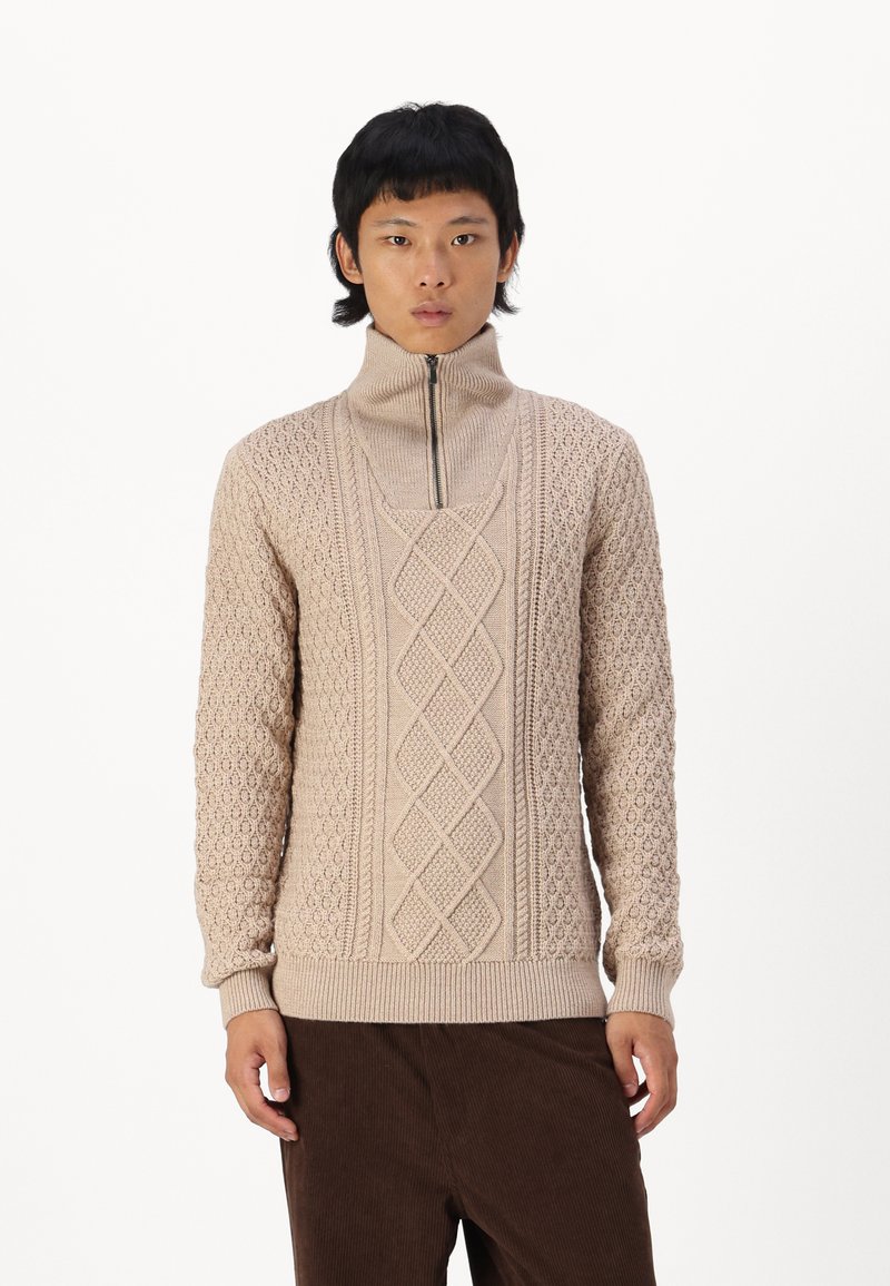Pull en tricot beige avec col montant, fermeture éclair, motifs texturés avec câbles et cercles, et poignets et ourlet côtelés.