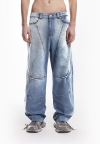 Svijetloplave traper cargo hlače s slobodnim krojem, koje sadrže jedinstvene zakrivljene šavove i istrošenu teksturu. Uparene s sivim tenisicama.