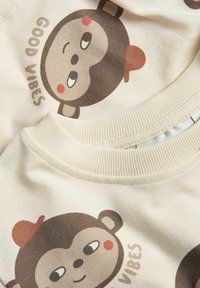 Krämfärgad tyg med en lekfull apa-illustration med röd hatt och texten "GOOD VIBES." Ribbad halsringning med mjuk textur.