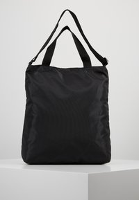 Musta nailoninen kassi, jossa on kaksinkertaiset lyhyet kahvat ja yksi pitkä säädettävä hihna, seisoo pystyssä valkoisella jalustalla yksinkertaista taustaa vasten.