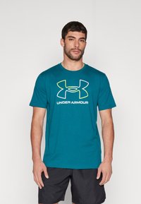 Mand iført en teal Under Armour t-shirt med logo og sorte shorts, stående mod en ensfarvet hvid baggrund.