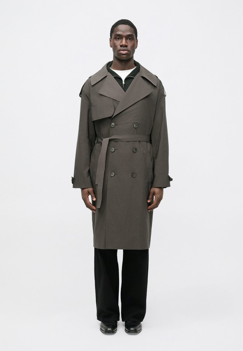 Dark gray trench coat with a double-breasted design, wide collars, and a waist tie, worn over a black knit top and trousers.