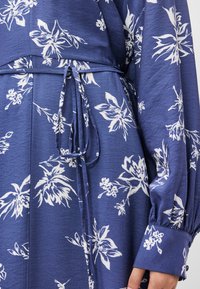 Μπλε φλοράλ φόρεμα με λευκά φλοράλ σχέδια, ζωνάκι για δέσιμο στη μέση και μακριά φουσκωτά μανίκια. Το ύφασμα φαίνεται μαλακό με ελαφριά υφή.