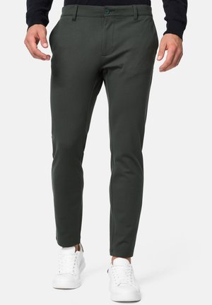 Dark green stretch pants with a slim fit design, featuring a button closure, side pockets, and paired with white sneakers. Smooth texture.