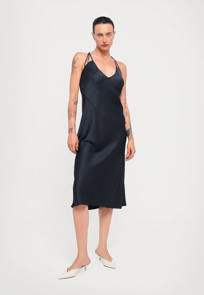 Marineblauw satijnen slipdress met een V-hals, gekruiste banden en een ruitpatroon. De lengte valt onder de knie en is gecombineerd met crèmekleurige muiltjes.