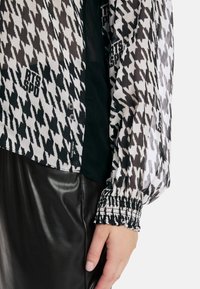 Schwarz-weißes Hahnentrittmuster-Bluse aus durchscheinendem Stoff, mit elastischen Bündchen und kleinen Knöpfen. Kombiniert mit einem glänzenden schwarzen Lederrock.