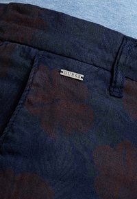 Shorts navy a fiori realizzati in tessuto leggero, con motivi floreali marrone scuro e una piccola etichetta argentata "GUESS" sulla vita.