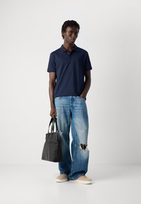 Férfi sötétkék pólót, világoskék, szakadt, bő szárú farmernadrágot, bézs sportcipőt visel, fekete vászontáskával a kezében, egy világos, egyszínű háttér előtt áll.