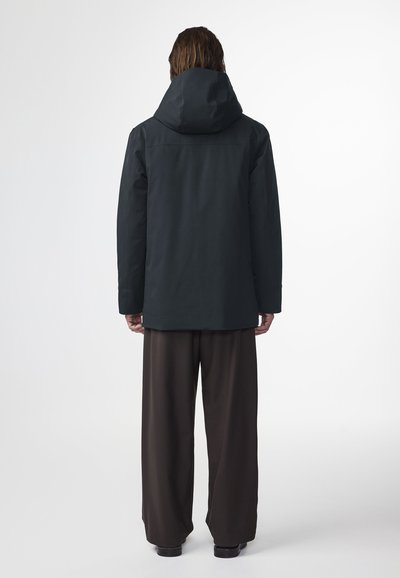 Giacca nera impermeabile con un grande cappuccio, taglio dritto e texture liscia. Indossata con pantaloni marroni larghi. Design minimalista.