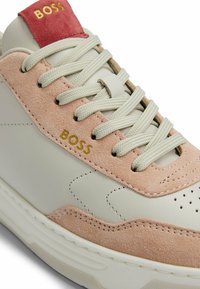 Sneaker in pelle bianca con accenti in suede rosa chiaro, logo "BOSS" in oro, lacci beige e dettagli perforati sulla punta. Materiali testurizzati.