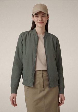 Kobieta w beżowej czapce, jasnobeżowej koszuli, oliwkowej kurtce zapinanej na zamek i khaki spódnicy, stoi na tle jednolitego jasnego tła.