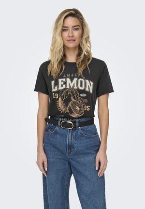 Joven con cabello largo y rubio que lleva una camiseta negra con estampado de limón y jeans azules de talle alto con cinturón negro sobre un fondo liso.