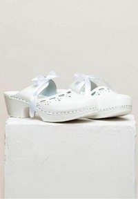 Witte klompen met uitsnijdingen, kleine strikjes en een platformhak. Gladde leren textuur met contrasterende stiksels langs de zool.