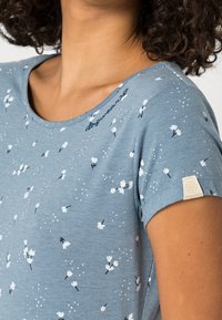 Jasnoniebieska koszulka z białym, kwiatowym nadrukiem na całej powierzchni, okrągłym dekoltem i krótkimi rękawami. Na lewym rękawie znajduje się metka z logo.