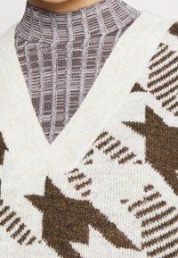 Kremowy sweter z dzianiny z dekoltem w serek, brązowymi wzorami gwiazd i teksturowanymi akcentami. Noszony na dopasowanym, w paski, szarym topie z wysokim kołnierzem.
