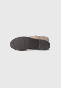Beige suède laars met een gestructureerde zwarte rubberen zool met een sterrenpatroon. Het bovenste deel is glad met stiksels langs de zijkanten.