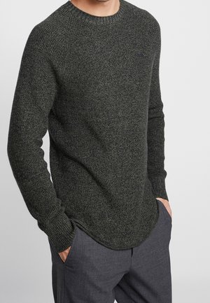 Uomo che indossa un maglione a maglia grigio scuro con maniche lunghe e tessuto strutturato, abbinato a pantaloni grigio scuro, con le mani nelle tasche.