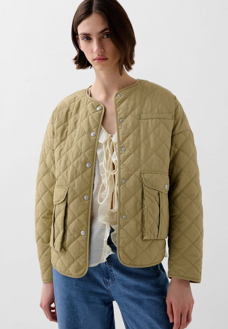 Vatteret khaki jakke med trykknapper, rund halsudskæring og forlommer. Bæres over en lys, flæset top med løse denim jeans.