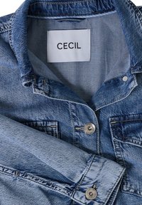 Blauwe spijkerjack met zilveren knopen, opgerolde mouw en een wit kledinglabel met de tekst "CECIL" binnenin de kraag.